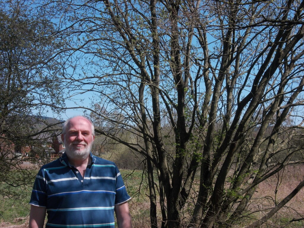An older man with a white beard and short hair - John Knight - is standing outdoors in front of a cluster of leafless and sparsely budding trees on Danes Moss on a sunny day. He is wearing a blue and green striped polo shirt and smiling at the camera. In the background, there is a grassy field, a few red-brick buildings, and hills under a clear blue sky.